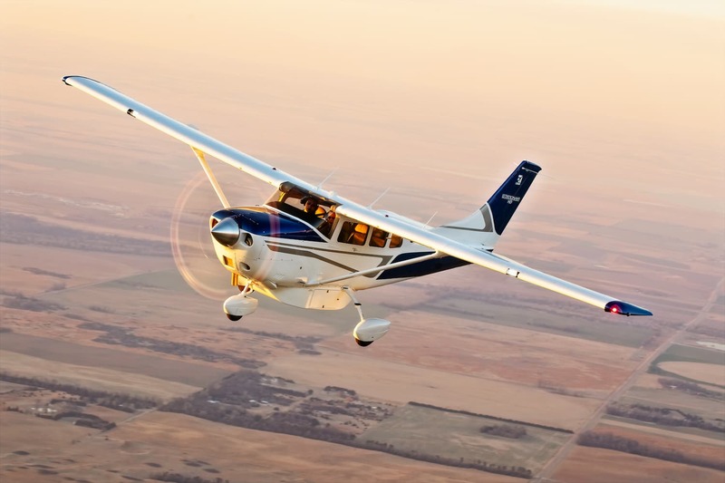 Imagem de um avião a pistão Cessna T206H Turbo Stationair HD branco e azul sobrevoando região rural.